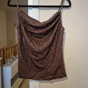 Brun glittrig linnetopp - Säljer en brun glittrig linnetopp med tunna axelband och draperad urringning. Toppen har ett skimrande material med små färgglada paljetter som ger en festlig känsla.