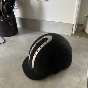 Svart ridhjälm från CRW - Säljer en svart ridhjälm från CRW med snygga silvriga detaljer på ovansidan. Hjälmen har klassisk form och passar perfekt för ridning. Märket är Classic Riding Wear och storleken är 57. Knappt använd typ 10 gånger men inga skador på den. Kostade runt 1000 kronor ny.