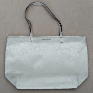 Silverfärgad shoppingväska från Victoria's Secret - Säljer en snygg och rymlig shoppingväska från Victoria's Secret i metallic silver. Väskan har två axelband och stängs med dragkedja. Perfekt för att bära med sig allt man behöver under dagen. Väskan har aldrig använts 