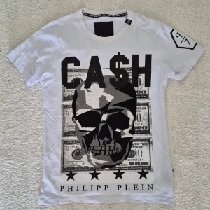 Philipp plein Vit t-shirt - Säljer en vit t-shirt från Philipp Plein med stort grafiskt tryck framtill. Motivet består av en döskalle, dollar-sedlar och texten 'CA$H'. T-shirten har rund hals och korta ärmar. Perfekt statement-plagg för dig som gillar streetstyle. I storlek M, i mycket fint skick. 