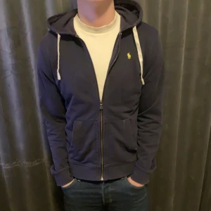 Marinblå hoodie från Ralph Lauren - Säljer en stilren marinblå hoodie från Ralph Lauren med dragkedja och vita snören i huvan. Den har en liten logga på bröstet och är perfekt för en avslappnad stil. Passar bra till jeans eller joggers.