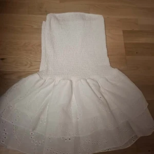 Vit kort klänning med volang - Supersöt vit klänning med smockad överdel och volangig kjol med broderade detaljer. Perfekt för dig som gillar romantisk och feminin stil. Klänningen är ärmlös och har en kort längd som ger en fräsch vibe. PRIS KAN GÅ NER