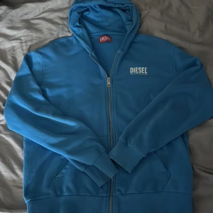 Blå hoodie från Diesel med dragkedja - Snygg blå hoodie från Diesel med vit logga på bröstet. Tröjan har huva, långa ärmar och praktisk dragkedja framtill. Ribbstickade muddar vid ärmslut och nederkant. Perfekt för chill dagar och streetstyle. Materialet är mjuk bomull som känns skönt mot huden.