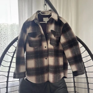 Rutig overshirt från H&M i S - Snygg rutig overshirt från H&M i storlek S. Färgerna är beige, brun och svart med stora rutmönster. Skjortan har två bröstfickor med lock och knappar framtill. Perfekt att slänga över en hoodie för en chill look.