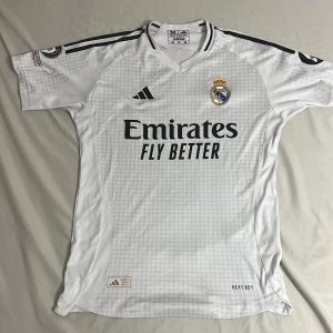 Real Madrid Mbappé 9 vit matchtröja - Säljer en officiell Real Madrid fotbollströja från Adidas i storlek M. Tröjan är vit med mörkblå detaljer, har korta ärmar och ett subtilt rutmönster. På framsidan finns klubbmärket och Emirates-sponsorn, på ryggen står Mbappé och nummer 9. Materialet är lätt och andas.