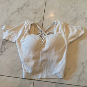 Beige croppad ribbad topp med snörning - Säljer en beige croppad topp med ribbad struktur och snygg snörning framtill. Toppen har korta ärmar, v-ringad hals och omlottkänsla med långa knytband som ger en cool look. Perfekt för dig som gillar trendiga detaljer och vill sticka ut.