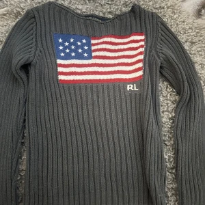 Grå stickad tröja Ralph Lauren M - Säljer en ribbstickad grå tröja från Ralph Lauren i storlek M, skulle säga att den är i XS-S mer!!!!!!!. Tröjan har ett stort amerikanskt flaggmotiv i rött, vitt och blått på bröstet samt RL-broderi. Långärmad och med en loose passform, perfekt för dig som gillar klassisk stil med en twist.