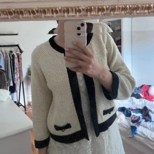 Beige och svart cardigan  - Snygg beige cardigan jacka med svarta detaljer och dekorativa knappar på fickorna. Jackan är stickad med grov struktur och har svarta kanter vid hals, ärmslut och nederkant. Perfekt för dig som gillar klassisk och chic stil.