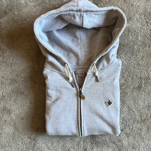 Ljusgrå hoodie med dragkedja Morris - Ljusgrå hoodie från Morris med dragkedja framtill och snörning i huvan. Tröjan har en broderad detalj på bröstet och ribbade muddar vid ärmslut och nederkant. Du får även med ett pärlarmband vid köpet! Storleken är M men det är en liten modell och passar S 
