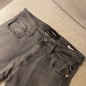 Replay Anbass grå jeans, storlek 31 - Snygga grå jeans från Replay, modell Anbass. Klassisk femficksdesign med Replay-logga på fickan och svart patch bak. Jeansen har en rak passform och är tillverkade i mjukt jeanstyg som sitter skönt hela dagen. Perfekt för dig som gillar stilrena och moderna jeans.