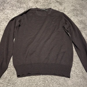 Mörkbrun stickad tröja J.Lindeberg - Snygg mörkbrun stickad tröja från J.Lindeberg i storlek M. Tröjan har rund halsringning och långa ärmar, med ribbade muddar vid ärmslut och nederkant. Perfekt för dig som gillar stilrena och enkla plagg med premiumkänsla.
