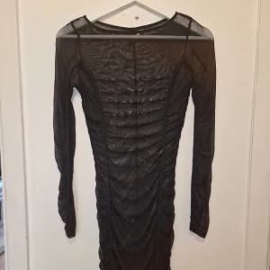 Svart meshklänning med rynkade detaljer - Svart meshklänning med långa ärmar och snygga rynkade detaljer längs hela klänningen. Den har en transparent look och snygg över en outfit.