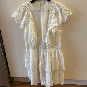 Kortklänning med volanger från H&M - Supersöt vit klänning från H&M med broderade volanger och kort ärm. Klänningen har omlottvolanger längs axlarna och flera lager med broderade detaljer. Perfekt för sommarens alla tillfällen, med elastisk midja och lätt, luftig bomull.