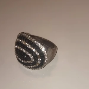 Glittrig silverring med svarta stenar - Säljer en chunky silverring med glittriga rader av små vita och svarta stenar i ett coolt, böljande mönster. Ringen har en bred form och sticker verkligen ut med sin blingiga look. Perfekt för dig som vill ha något som syns och känns extra.