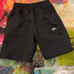 Svarta shorts från Lacoste - Svarta shorts från Lacoste med klassisk krokodillogga broderad på benet. De har elastisk midja och två sidofickor, vilket gör dem riktigt sköna och praktiska. Perfekta för chill dagar eller sport. Materialet känns mjukt och är troligtvis bomull.är väldigt små för mig passar xxs och ungefär 145cm 140 skulle jag säga 