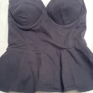 Svart korsett peplumtop - Svart korsett peplumtop med markerade kupor och figurnära passform. Toppen är axelbandslös och har en snygg volang nedtill som ger extra stilpoäng. Perfekt för dig som vill ha en edgy och trendig look.