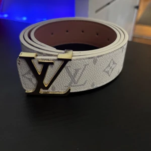 Vit Louis Vuitton skärp med LV-spänne - Säljer ett stilrent vitt Louis Vuitton bälte med ikoniskt LV-spänne i guld. Bältet har klassiskt monogrammönster i ljusgrått och är tillverkat i exklusivt skinn. Perfekt accessoar för dig som vill ha en lyxig och trendig look.