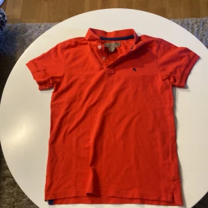 Röd pikétröja från L.O.G.G. - Snygg röd pikétröja från L.O.G.G. med klassisk krage och korta ärmar. Tröjan har knappar vid halsen och ett mörkblått broderat djur på bröstet. Tillverkad i mjuk bomull som är skön mot huden. Perfekt för dig som gillar stilren och enkel design.