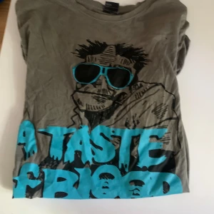 Grå t-shirt med tryck och blå text - Säljer en grå t-shirt från H&M med coolt tryck av en tecknad person med blå solglasögon och stor blå text. T-shirten har rund hals och korta ärmar, perfekt för dig som gillar grafiska motiv och streetstyle.