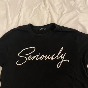 Svart croppad t-shirt FB Sister - Svart croppad t-shirt från FB Sister med vit broderad text 'Seriously' på framsidan. T-shirten har rund hals och korta ärmar, tillverkad i mjuk bomull. Perfekt för en chill och avslappnad stil.
