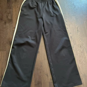 Svarta wide byxor med vit/gul rand - Svarta wide byxor med elastisk midja och breda ben. Snygg vit rand med gul kant längs sidorna ger en sportig vibe. Tillverkade i mjukt och stretchigt material, 94% polyester och 6% elastan. Perfekta för chill eller träning.