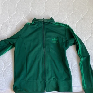 Grön Adidas track jacket L - Snygg grön Adidas track jacket med klassiska tre ränder längs ärmarna och Adidas logga på bröstet. Jackan har hel dragkedja, hög krage och är tillverkad i mjuk polyester. Perfekt för en sportig streetstyle-look.