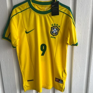 Brasilien Ronaldo 9 fotbollströja Nike - Säljer en ikonisk Brasilien fotbollströja från Nike med Ronaldo och nummer 9 på ryggen. Tröjan är gul med gröna detaljer, korta ärmar och rund hals. Officiell CBF-logga och Nike Swoosh på bröstet. Tillverkad i lätt och ventilerande polyester.