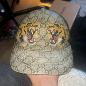 Gucci keps XS med tigertryck - Säljer en Gucci keps i storlek XS med ikoniskt GG-mönster i beige och brunt, prydd med två stora tigerhuvuden framtill. Baksidan har svart mesh och justerbart band i grönt och rött. Perfekt för dig som vill sticka ut med lyxig streetstyle.