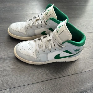 Nike Air Jordan 1 Mid vit/grön EUR 39 junior  - Nike Air Jordan 1 Mid sneakers i vitt och grönt med klassisk Jordan-logga på plösen och sidan. Skorna har snörning, perforerad tå och en grön insida som poppar mot det vita skinnet. Snygg retrostil med rund tå och platt sula, perfekt för dig som gillar streetwear.                                       Använd endast en kortare period.