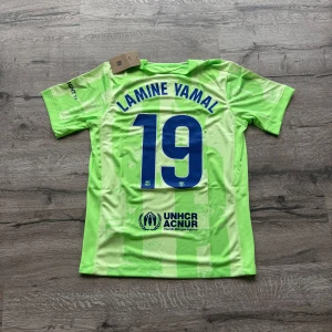 Barcelona Lamine Yamal #19 fotbollströja - Snygg Barcelona fotbollströja med Lamine Yamal och nummer 19 på ryggen. Tröjan är ljusgrön med subtilt randigt mönster, blå tryck och klubbmärke på bröstet. Materialet är lätt och andas, perfekt för match eller träning. Sponsorer som Spotify och UNHCR syns tydligt. Storlek: S helt ny med prislapp. Priset är inte hugget i sten så skicka pm vid funderingar.