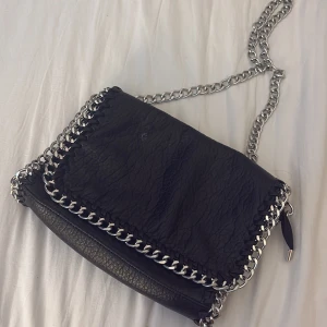 Svart axelväska med kedjedetaljer från Tiamo - Snygg svart axelväska från Tiamo i skinn med grova silverfärgade kedjor runt kanterna och som axelrem. Väskan har en dragkedja på baksidan och flera fack inuti. Perfekt för dig som gillar en edgy stil och vill ha plats för allt viktigt.