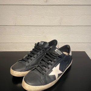 Svarta Golden Goose sneakers - Golden Goose sneakers i svart skinn med vit stjärndetalj på sidan och vit häl. Klassisk låg modell med rund tå, svarta snören och platt sula. Märkets logga på plösen. Levereras med originalpåse och box. Sulan har släppt lite bak i hälen därav priset