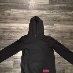 Svart hoodie från Calvin Klein - Svart hoodie från Calvin Klein med lång ärm och huva. Tröjan har en röd logotyp-patch framtill och en klassisk känguruficka. Materialet är mjuk bomull och passformen är normal. Perfekt för chill dagar och streetwear-stil.