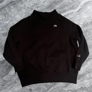 Svart sweatshirt från Champion - Svart sweatshirt från Champion med liten logga på bröstet och ärmen. Tröjan har en hög krage och långa ärmar med breda muddar. Materialet är mjuk bomull och passformen är normal. Perfekt för chill dagar eller när du vill ha en clean look.