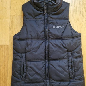 Svart dunväst från H&M strl 152 - Säljer en svart dunväst från H&M med broderad SNW-7-logga på bröstet. Västen har hög krage, två fickor framtill och stängs med dragkedja. Perfekt för lager-på-lager och enkel att matcha med hoodies eller tröjor.