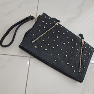 Svart kuvertväska med nitar och dragkedjor - Snygg svart kuvertväska med guldfärgade nitar och två dragkedjor på framsidan. Väskan har en handledsrem och är tillverkad i skinnimitation. Perfekt för att ge din outfit lite edge och att bära det viktigaste.