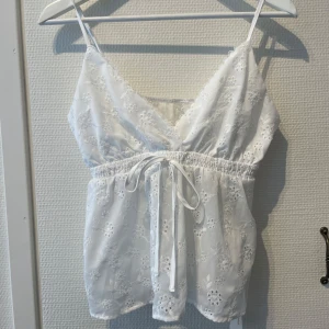 Vit broderad linnetopp med roset - Superfin vit linnetopp med smala axelband, v-ringad hals och broderade blommor över hela tyget. Toppen har spetsdetaljer längs halsen och en söt knytning i midjan som ger en feminin siluett. Perfekt för varma dagar och sommarhäng. Aldrig använd!❤️ 