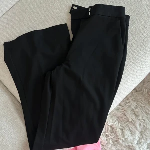 Svarta kostymbyxor med raka ben - Snygga svarta kostymbyxor med klassisk rak passform och diskreta fickor bak. Byxorna har en stilren midja med hyska och hake, perfekt för en clean och dressad look. Materialet känns mjukt och har en lätt stretch för extra komfort.