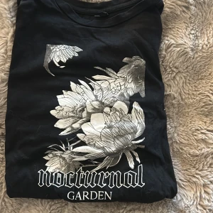 Svart t-shirt Nocturnal Garden S - Svart t-shirt i storlek S med silvrigt blommigt tryck och texten 'nocturnal garden' framtill. Rund hals och klassisk passform. Perfekt för dig som gillar coola prints och lite mystisk vibe. Materialet känns mjukt och skönt mot huden.