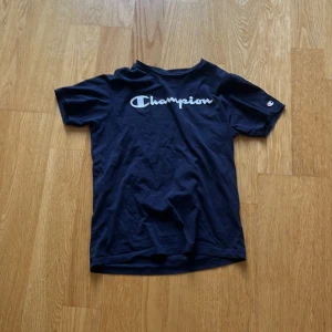 Mörkblå Champion t-shirt X-Large - Snygg mörkblå t-shirt från Champion i storlek X-Large barn. Klassisk modell med korta ärmar och rund hals. Stor Champion-logga i vitt på bröstet och liten logga på ärmen. Tillverkad i mjuk bomull som är skön mot huden.
