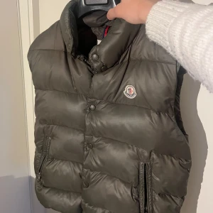 Mörkgrå dunväst från Moncler - Snygg mörkgrön dunväst från Moncler med hög krage och ikonisk logga på bröstet. Västen har quiltad design, tryckknappar och dragkedjor vid fickorna. Size 3/M