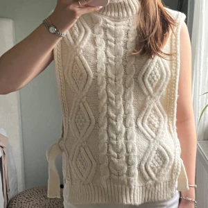 Stickad vit väst med snörning - Säljer en chunky stickad väst i vit färg med grova flätstickade detaljer och diamantmönster. Västen har hög hals och snörning vid sidorna!🫶🏻