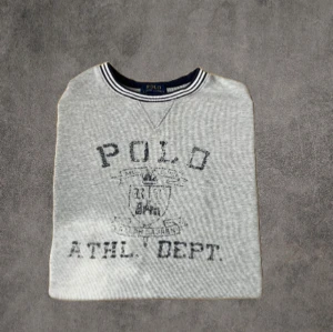 Grå sweatshirt från Polo Ralph Lauren - Grå sweatshirt från Polo Ralph Lauren med tryckt logga och text framtill. Tröjan har rund hals med randig ribbad kant i vitt och marinblått. Klassisk och enkel stil, perfekt till jeans eller joggers. Mjuk bomullsblandning för skön känsla.