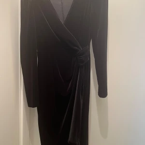 Svart omlottklänning i sammet - Snygg svart långklänning i mjuk sammet med omlott-design och knyt i midjan. Klänningen har långa ärmar och en elegant v-ringad hals. Perfekt för dig som vill ha en stilren och lyxig look med lite extra edge.