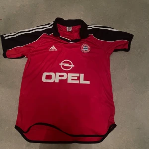 Bayern München röd Adidas fotbollströja - Säljer en retro Bayern München fotbollströja från 2002, Adidas i rött med mörkblått och vita detaljer. Tröjan har korta ärmar, vit OPEL-logga på bröstet och klubbmärke. Materialet är lätt och andas, perfekt för match eller träning. Snygga vita ränder på axlarna och Bayern München-tryck på ryggen. Har ni funderingar så är de bara och fråga!