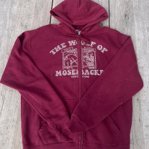 Vinröd hoodie från One of One - Säljer en vinröd hoodie från One of One i storlek medium. Tröjan har huva med snörning, dragkedja framtill och ett tryck med texten 'The Wolf of Mosebacke' i vita prickar. Mjuk insida och två fickor fram. Perfekt för dig som gillar streetwear vibes.