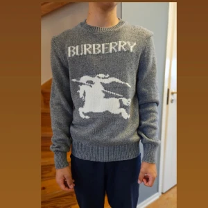 Grå stickad tröja från Burberry - Säljer en stilren grå stickad tröja från Burberry med ett stort vitt logotypmotiv på framsidan. Tröjan har långa ärmar och en rund halsringning med ribbade kanter.  Oanvänd. Kvitto saknas.