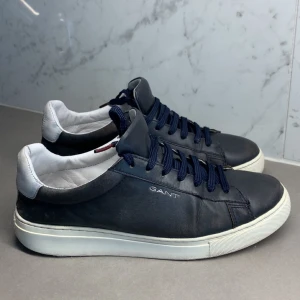 Mörkblå sneakers från GANT - Snygga mörkblå sneakers från GANT med vit sula och vita detaljer vid hälen. Skorna har rund tå, platt sula och klassisk snörning med mörkblå skosnören. Tillverkade i skinn för en stilren och clean look. Perfekta för dig som gillar minimalistisk design.
