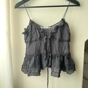 svart volangtop från Zara med snörning - Supersöt grå linnetopp från Zara med tunna axelband, volanger och snörning framtill. Toppen har en luftig och flowy passform med flera lager volangdetaljer och en lätt skrynklig struktur. Perfekt för att ge din outfit en romantisk vibe.
