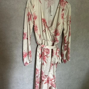 Blommig omlottklänning från Gina Tricot - Säljer en vacker lång omlottklänning från Gina Tricot i ljus nyans med rosa blommigt mönster. Klänningen har lång ärm, v-ringning och knytband i midjan för en smickrande passform. Perfekt för sommarens tillfällen!